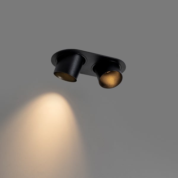 Schwarzer zweiflammiger Deckenstrahler mit zylindrischen Spotköpfen, eine Lichtquelle ist eingeschaltet und wirft warmes Licht.