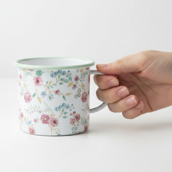 Emaille-Becher mit Blumenmuster und grünem Rand, von einer Hand gehalten.