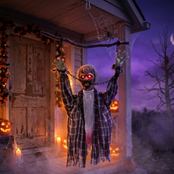 Hängende Halloween Dekofigur Zombie mit leuchtenden Augen an einem Ast, vor einem Haus mit Kürbissen und Spinnweben.