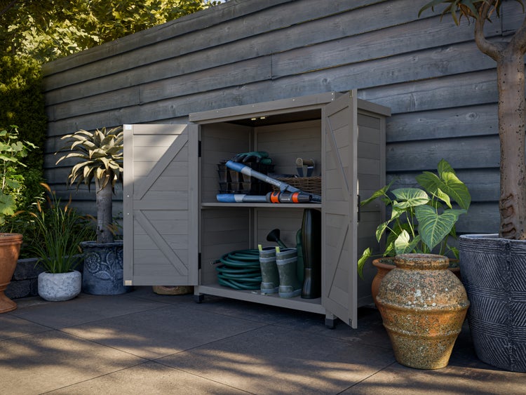 Gartenschrank aus Kunststoff in Holzoptik mit geöffneten Türen, gefüllt mit Gartengeräten und Zubehör auf einer Terrasse vor einem Holzzaun.