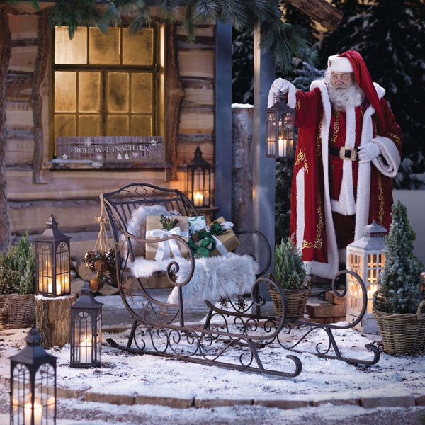 Weihnachtsszene mit Weihnachtsmann, Schlitten, Laternen und Geschenken vor einer Holzhütte