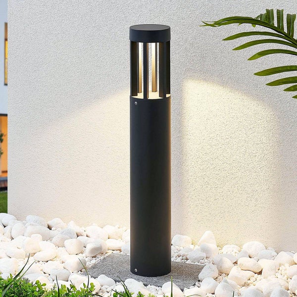 Moderne zylindrische Wegeleuchte in Schwarz, platziert auf weißen Kieselsteinen vor einer hellen Wand.