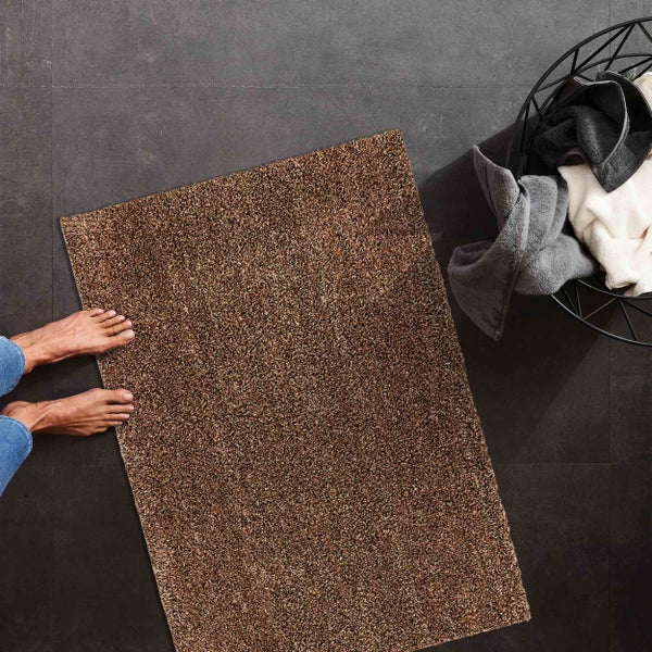 Rechteckige Hochflor-Badematte in Braun auf Bodenfliesen in einem Badezimmer neben einem Korb mit Handtüchern.