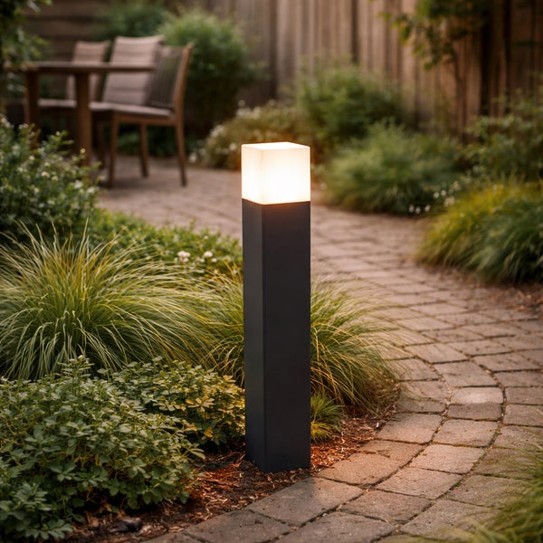 Moderne anthrazitfarbene Pollerleuchte mit quadratischem Diffusor an einem gepflasterten Gartenweg inmitten grüner Pflanzen.