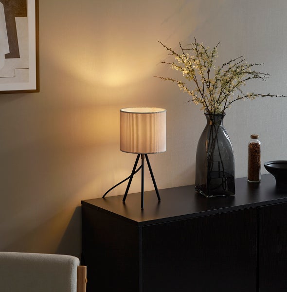 Moderne Dreibein-Tischleuchte mit zylindrischem Textilschirm auf einem Sideboard neben einer Glasvase mit Zweigen und Blumendekoration.