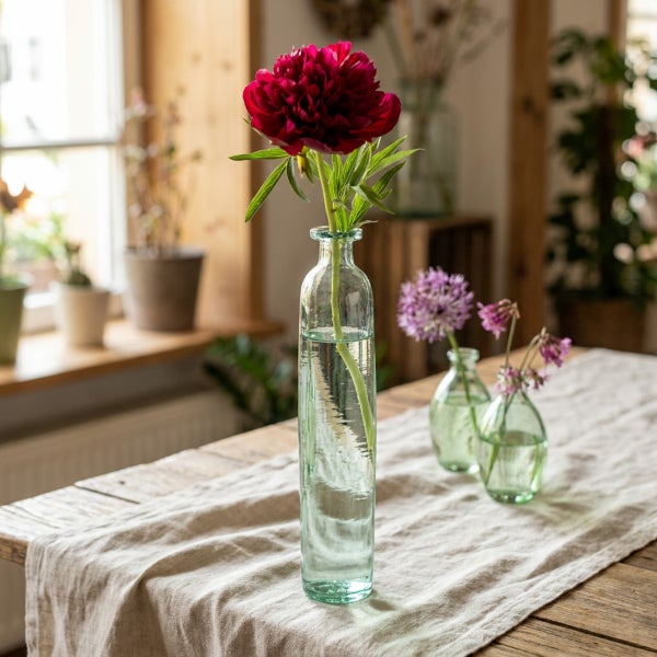 Drei kleine Glasvasen mit frischen Blumen stehen als Tischdekoration auf einem Holztisch mit einem beigen Tischläufer in einem hellen Raum.