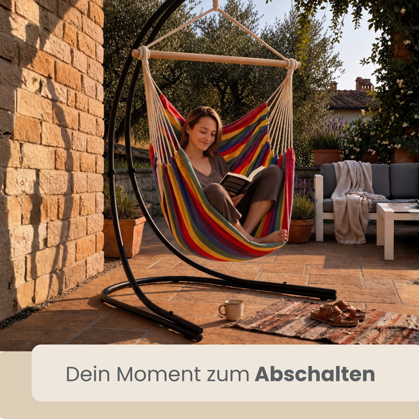Eine Frau liest in einer gestreiften Hängematte mit Gestell auf einer sonnigen Terrasse. Gartenmöbel und Backsteinmauer im Hintergrund.