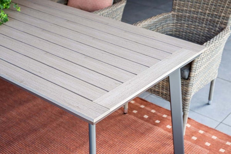 Gartentisch mit Tischplatte in Holzoptik und Metallgestell sowie Gartenstühle aus Geflecht auf einer Terrasse mit Außenteppich.
