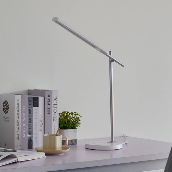 Moderne, minimalistische Schreibtischlampe mit schlankem weißem Kopf und silbernem Gestell auf einem organisierten Schreibtisch neben Büchern und einer Pflanze.