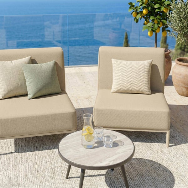 Moderne Gartenlounge mit zwei beigen Polstersesseln, Dekokissen, rundem Beistelltisch und Teppich auf einer Terrasse mit Meerblick.