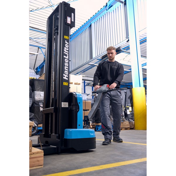 Eine Person bedient einen elektrischen HanseLifter Hochhubwagen in einer geräumigen Industriehalle mit Lagerregalen.