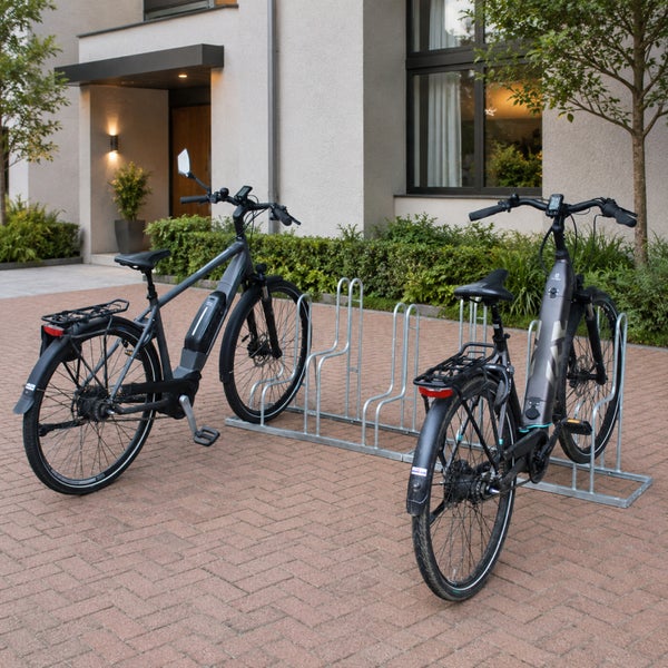 Zwei E-Bikes stehen in einem verzinkten Fahrradständer vor einem modernen Wohnhaus mit gepflastertem Vorplatz.