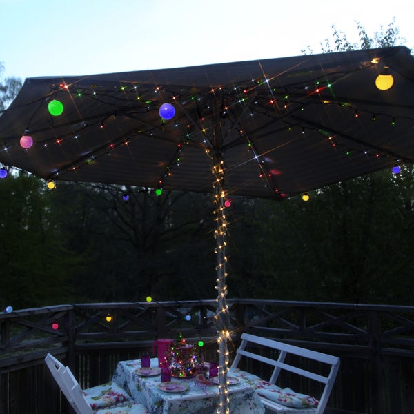 Szene mit einem Sonnenschirm, der mit bunten Lichtern und einer Lichterkette dekoriert ist, auf einer Terrasse mit Tisch und Stühlen