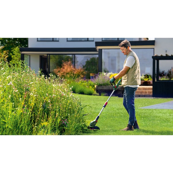 Ein Mann nutzt einen Akku Rasentrimmer für die Rasenkantenpflege im Garten vor einem modernen Wohnhaus.
