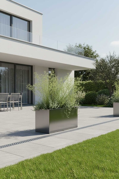 Rechteckiger Pflanzkübel aus Edelstahl mit Gräsern auf einer modernen Terrasse vor einem weißen Wohnhaus.