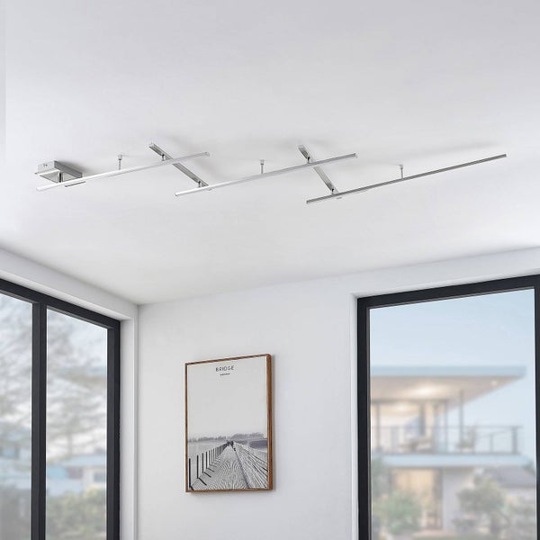 Moderne LED-Deckenleuchte aus verchromtem Metall mit versetzten Lichtbalken in einem hellen, minimalistischen Raum.