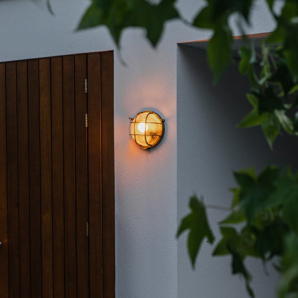 Runde Außenwandleuchte mit Schutzgitter und geriffeltem Glas an einer Wand neben einer Holztür.
