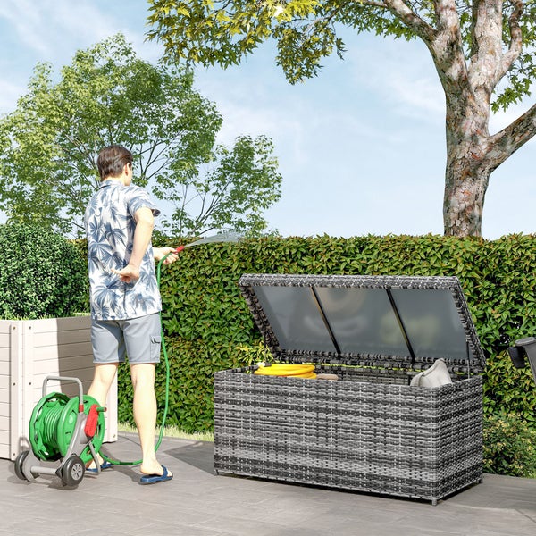 Szene im Garten mit einer offenen Gartenbox aus Rattan, einem Schlauchwagen und einer Person, die mit einem Gartenschlauch gießt
