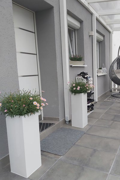 Moderner Terrassenbereich mit hohen weißen Pflanzsäulen, rosa Blumen, grauem Fliesenboden, Hauseingang und grauem Hängesessel.