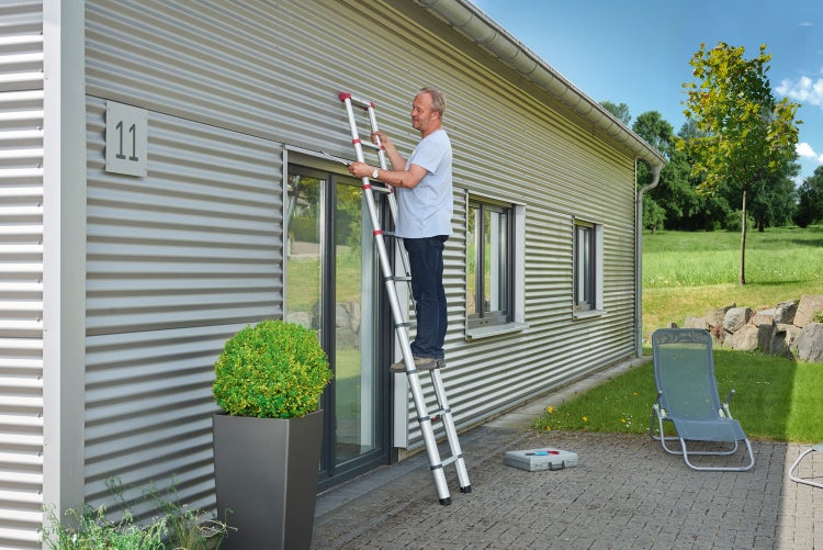 Mann steht auf einer Teleskopleiter vor einem Haus mit Wellblechfassade.