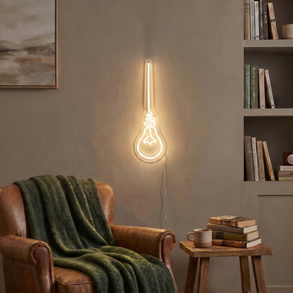 Wandleuchte in Form einer Glühbirne als Neon Lichtobjekt montiert über einem Ledersessel in einem gemütlichen Wohnzimmer mit Bücherregal.