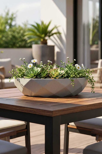 Moderner geometrischer Pflanztopf aus Stein mit Sukkulenten und weißen Blumen auf einem Holztisch im Außenbereich.