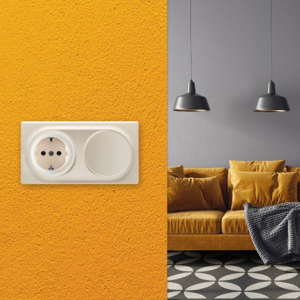 Eine Schuko Steckdose mit einem Blinddeckel auf einer strukturierten Wand in einem modernen Wohnzimmer mit Sofa und Hängeleuchten.