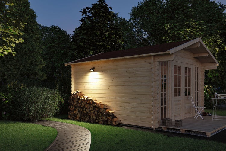 Holzgartenhaus bei Nacht mit Außenbeleuchtung, Kaminholzstapel, gepflastertem Weg und Bäumen.