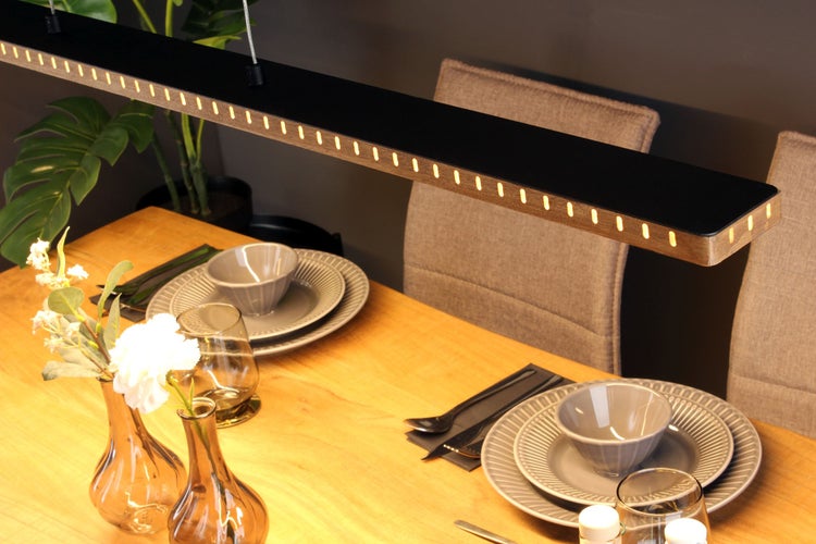 Moderne LED-Pendelleuchte aus Holz und Metall über einem gedeckten Esstisch aus Holz mit Tellern, Gläsern und Blumen in gemütlicher Atmosphäre.
