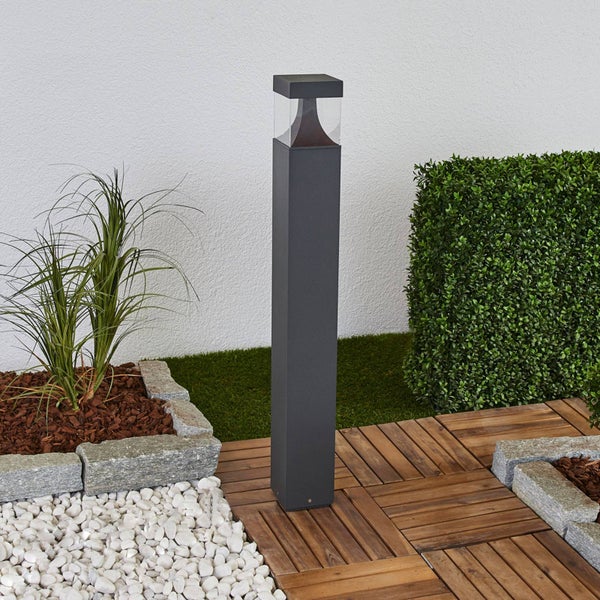 Moderne anthrazitfarbene Pollerleuchte in einem Garten mit Holzfliesen, weißen Steinen und einer Hecke vor einer weißen Wand.