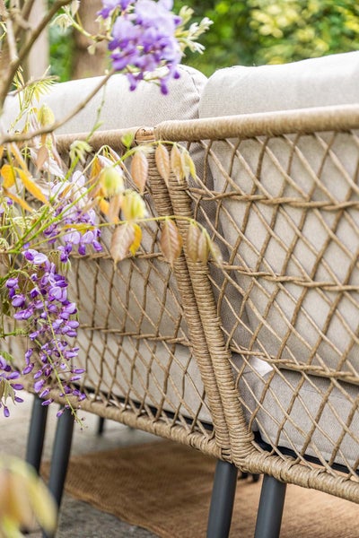 Detailaufnahme einer Garten-Lounge mit geflochtener Rückenlehne aus Seil und Polstern neben blühenden Pflanzen im Außenbereich.