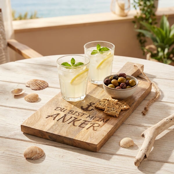 Servierbrett aus Holz mit der Gravur Du bist mein Anker, belegt mit zwei Gläsern Limonade, einer Schale Oliven und Gebäck auf einem Holztisch.
