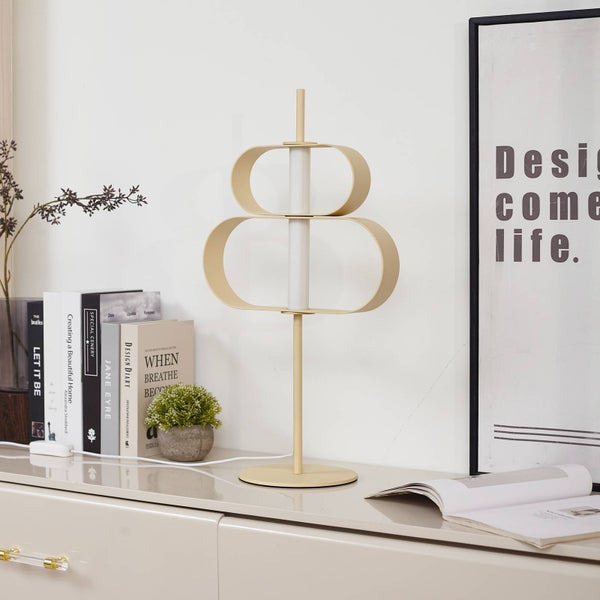 Moderne beige Tischleuchte aus Metall in skulpturalem Design auf einem Sideboard neben Büchern und einer kleinen Pflanze.