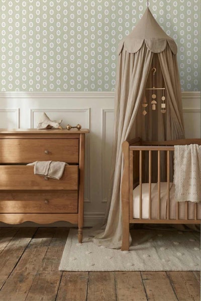 Kinderzimmer mit gemusterter Tapete, Wandvertäfelung, Holzkommode und einem Babybett mit Baldachin auf Holzfußboden.
