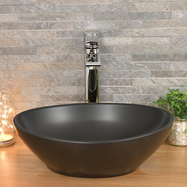 Rundes Aufsatzwaschbecken in Schwarz auf einer Holzplatte mit verchromter Armatur vor einer Wand mit Steinoptik Fliesen.