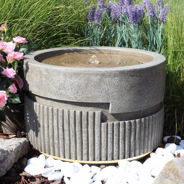 Moderner Gartenbrunnen in Steinoptik mit gerillter Struktur und sprudelndem Wasser in einer Gartenanlage mit Pflanzen und Zierkies.