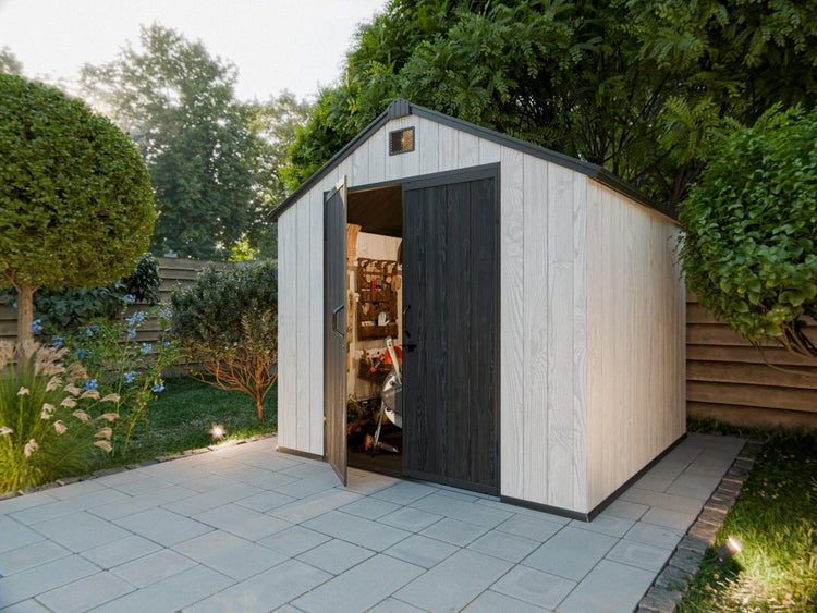 Modernes Gerätehaus in Holzoptik mit Doppeltür, eine Tür offen mit Blick auf Gartenwerkzeuge, auf einer gepflasterten Terrasse im Garten.