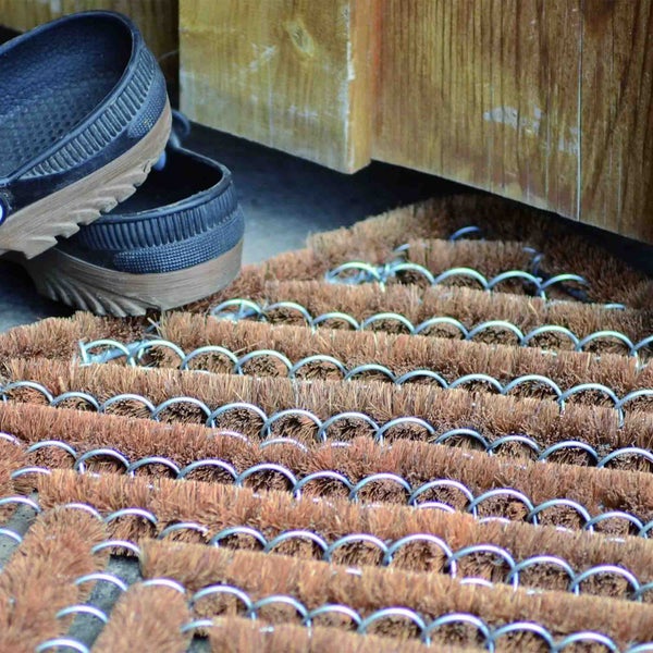 Fußmatte aus Kokosfasern mit Kettengliedern und Paar Clogs vor einer Holzwand