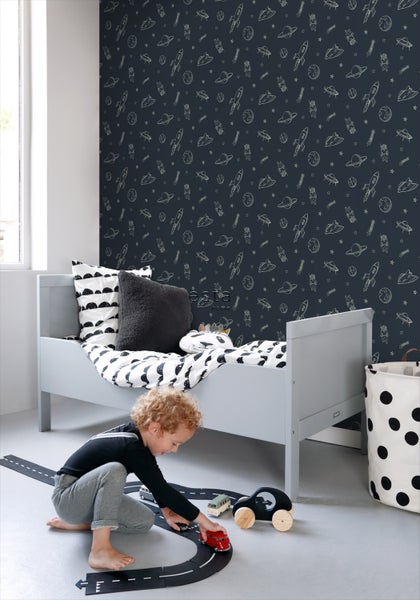 Kinderzimmer mit einem grauen Bett, einer Tapete mit Weltraummotiven und einem Jungen, der auf einem Teppich mit Autospielzeug spielt