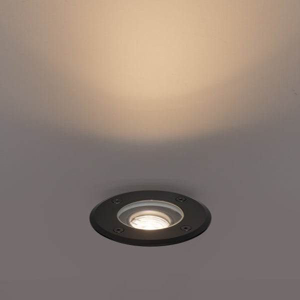 Ein runder Bodeneinbaustrahler in Schwarz mit warmweißem Lichtkegel auf einer grauen Fläche.