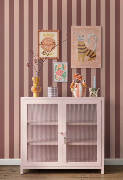 Rosa Metallschrank mit Riffelglastüren vor gestreifter Tapete, dekoriert mit Vasen und Wandbildern in einem modern gestalteten Raum.