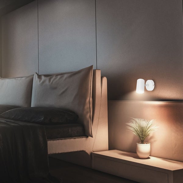 Weiße, zylindrische Wandleuchte in einem modernen Schlafzimmer, die eine kleine Zimmerpflanze auf einer Ablage neben dem Bett beleuchtet.
