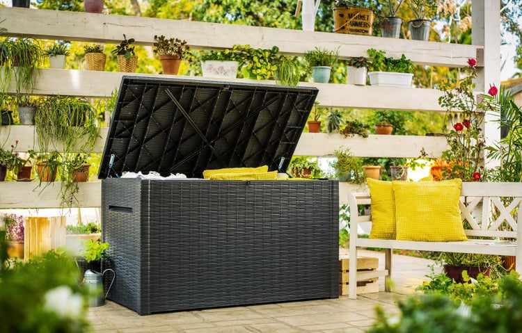 Eine geöffnete Gartenbox mit Rattanoptik auf einer Terrasse mit Gartenmöbeln und Pflanzen.