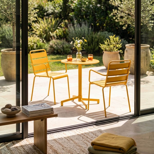 Runder, gelber Bistrotisch aus Metall auf einer hellen Steinterrasse mit Gartenhintergrund. Auf dem Tisch stehen zwei Gläser und eine Blumenvase.