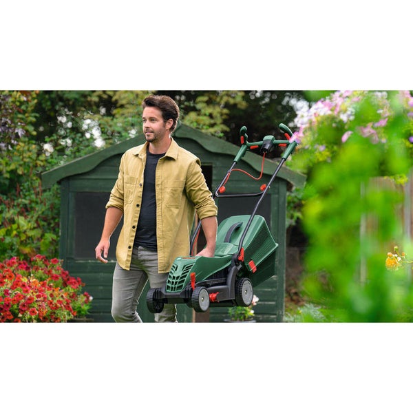 Ein Mann trägt einen kompakten grünen Elektro-Rasenmäher in einem hellen Garten mit Blumen vor einem Gartenhaus aus Holz.