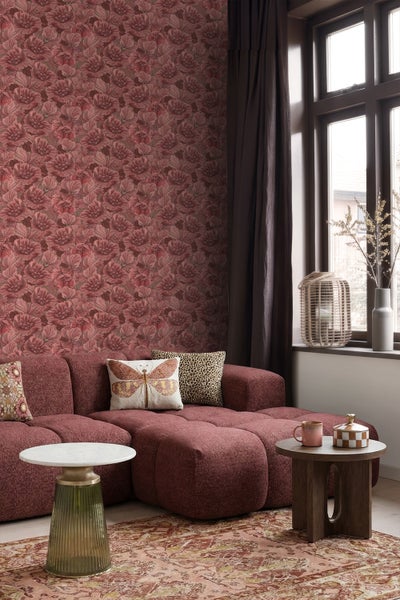 Vliestapete mit floralem Muster in Rottönen in einem Wohnzimmer mit bordeauxrotem Sofa und Beistelltischen.