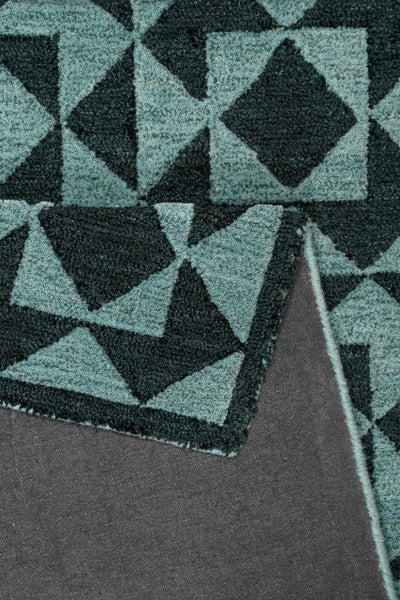 Türkisfarbener Kurzflor-Teppich mit geometrischem Muster aus Rauten und Dreiecken, umgeknickte Ecke zeigt graue, rutschfeste Rückseite.