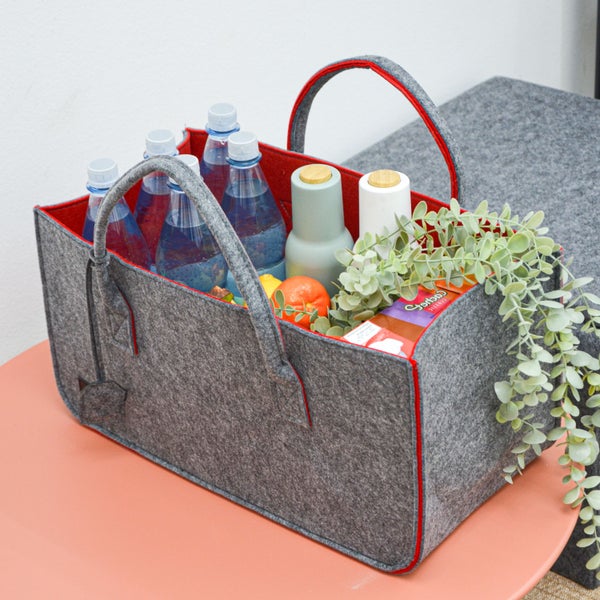 Graue Filztasche als Einkaufskorb gefüllt mit Getränkeflaschen, Obst und einer Pflanze, stehend auf einem runden Tisch