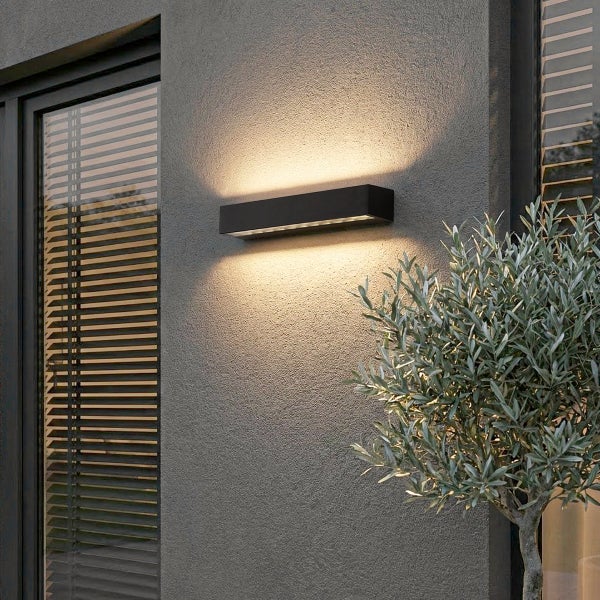Rechteckige schwarze LED-Außenwandleuchte an einer grauen Fassade mit Lichtschein nach oben und unten, neben Fenster und Pflanze.