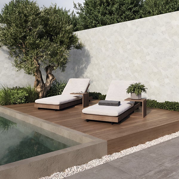 Zwei moderne Gartenliegen aus Holz mit hellen Auflagen stehen auf einer Holzterrasse am Pool vor einer hellen Steinwand und einem Olivenbaum.